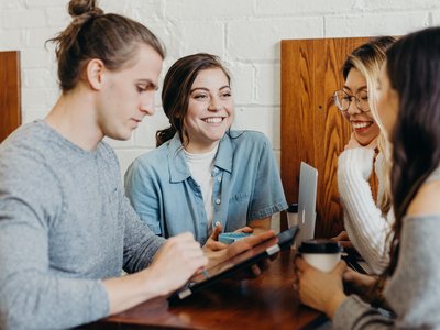 Drei junge Frauen und ein junger Mann unterhalten sich an einem Tisch; der Mann schaut auf einen Tablet Computer.